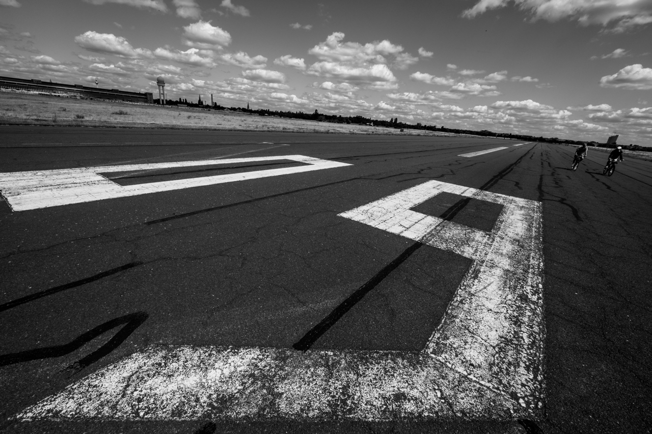 A black-and-white photo of one of Tempelhof's old runways - the '09' marker on the pavement in the foreground, two people biking on the runway in the background