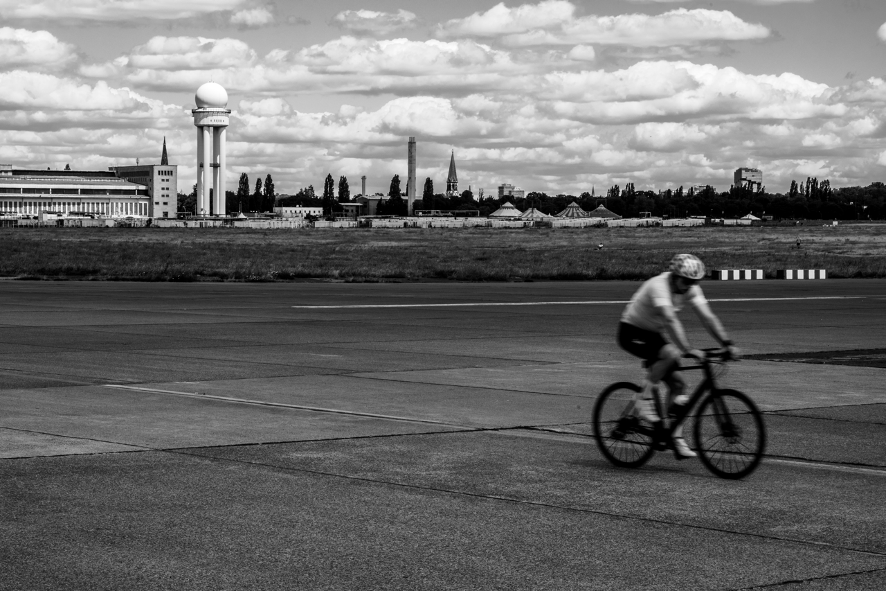 A black-and-white photo of a man biking along the concrete tarmac at Tempelhof.  The old radar dome is in the background.