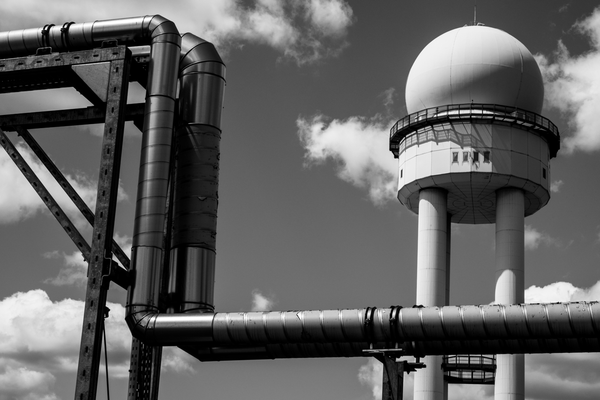 A black and white photo of the old radar dome at Tempelhof, Berlin, with some pipes in the foreground and partly-cloudy sky in the background